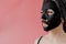 Young woman apply black cosmetic fabric facial mask on pink background. Face peeling mask with charcoal, spa beauty treatment,
