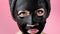 Young woman apply black cosmetic fabric facial mask on pink background. Face peeling mask with charcoal, spa beauty treatment