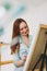 Young woman artist with long red hair draws picture with palette knife on easel