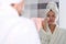 Young woman in bathrobe and towel on head washing off cosmetics from face in front of mirror