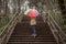 young woman in a beige coat and rain boots stands in forest or park and holds colorful bright rainbow umbrella