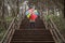 young woman in a beige coat and rain boots stands in forest or park and holds colorful bright rainbow umbrella