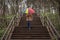 young woman in a beige coat and rain boots stands in forest or park and holds colorful bright rainbow umbrella