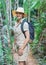 Young woman in beige shirt, shorts and straw haw, backpack on shoulders, smiling, jungle around