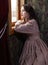 Young woman in beige vintage dress looking trough the window in