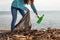 A young woman bends down to collect a dirty glass bottle. Cleaning and environmental protection