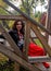 A young woman in a black leather jacket, red skirt and beads sitting and look smiling through a wooden staircase banister