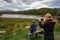 Young woman blogger takes a photo of her friend from behind against mountain lake. Tourist picnic on the background of beautiful