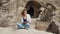 A young woman blonde in blue jeans sitting on a stone and looking at the phone. Background of the ancient city