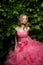 Young woman with blonde locks and makeup wearing pink evening dress with fluffy skirt is posing outdoors near the bush