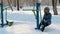 Young woman in blue down jacket with hood is doing legs exercises on simulator in a winter Park.