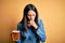 Young woman with blue eyes drinking jar of beer standing over isolated yellow background feeling unwell and coughing as symptom