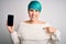 Young woman with blue fashion hair holding smartphone showing screen with surprise face pointing finger to himself