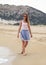 Young woman in blue skirt, t shirt and sunglasses walking on beach, smiling, overcast weather, hill behind her