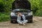 A young woman broke a car on the road and she sits in front of a car with an open hood in confusion and waiting for help with a ca