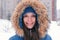 Young woman brunette looking at camera and smiling. Wearing blue down jacket with fur hood, face close-up.