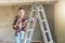 A young woman builder, dressed in a plaid shirt and construction glasses, stands in the workshop on a stepladder