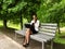 Young woman in a business suit drinks coffee and works on a laptop while sitting on a bench in the park, side view. Lonely adult