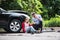 Young woman by the car after an accident and a man making a phone call.