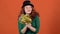 Young woman celebrating saint patrick`s day on orange wall in hat holding pom poms