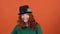 Young woman celebrating saint patrick`s day on orange wall in hat putting hair up