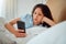 Young woman checking phone and social media in bed, browsing internet after waking up to text message at home. Female