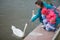 The young woman with the child feeds a white swan in a city pond with dandelions