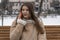 Young woman communicates by gadget. Brunette girl in sheepskin coat sits on bench in winter park and talks on smartphone