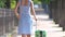Young woman commuter talking on mobile phone and going away on sidewalk pulling green suitcase walking down the street