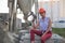 Young woman in construction helmet with drill sits on cement and concrete slabs on construction site near huge pile of sand