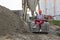 Young woman in construction helmet with drill sits on cement and concrete slabs on construction site near huge pile of sand