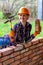 A young woman in a construction helmet with a hammer and a brick in her hands smiles and builds a wall of bricks.