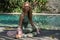 Young woman with curly long blonde hair and blue eyes sitting in Gomukhasana yoga pose next to blue swimming pool