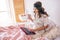 Young woman with dark hair Sitting on bed in bedroom alone. Holding cup and waoking. Plate with cakes on table. Remote