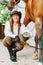 Young woman dressed in riding clothes cleaning the hooves of her brown horse in a stall