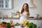 Young woman drinking green smoothie and fresh water with cucumber, lemon, leaves of mint on the kitchen table with fruits and