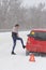 Young woman driver checks tire of  car on road in  blizzard and bad weather. Triangular accident warning sign stands on the