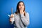 Young woman eating asian noodles from take away box using chopstick over blue background cover mouth with hand shocked with shame