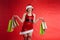 A young woman elf dressed as Santa in round glasses holds d background, hands holding a gift package, many paper bags from the