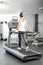 A young woman in an exercise room is running on an automatic treadmill.