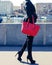 A young woman fashion blogger walks along the waterfront in a black trendy suit, high heels and with a large red handbag