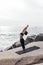 Young woman in fitness wear doing warming up exercises while standing on mat by seaside. Female practicing Hasta Uttanasana pose