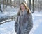 Young woman in a fur coat stands on the bank of a stream in a winter park