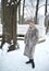 Young woman in a fur coat stands near a tree in a winter park