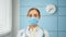 Young woman general practitioner in white coat and blue disposable face mask looks straight