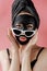 Young woman in glasses appling black cosmetic fabric facial mask on pink background. Face peeling mask with charcoal, spa beauty