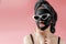 Young woman in glasses appling black cosmetic fabric facial mask on pink background. Face peeling mask with charcoal, spa beauty