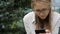 Young woman in glasses and office clothes is looking at the phone while sitting in the park on a bench.