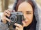 Young woman in gray coat photographing on old photo camera in forest. Pretty female taking photos with old camera in