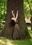 Young woman in green dress meditatively relaxes near large tree in forest park, concept of purity of nature and unity with